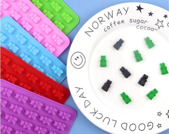 May include: Six silicone moulds for making gummy bears, each mould is a different colour: purple, red, blue, pink, green, and grey. The moulds are arranged in a row. The moulds are 18.5 cm by 13.5 cm by 1 cm. A plate with gummy bears in various colours is in the background.