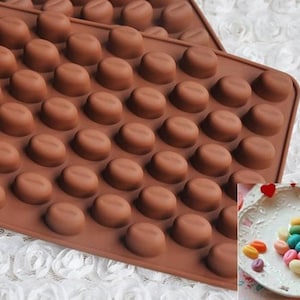 以下が含まれることがあります： コーヒー豆の形をした48個の個別のキャビティを持つ茶色のシリコンモールド。モールドは白いレースの表面にあります。背景には、コーヒー豆の形をしたカラフルなキャンディーが入ったプレートがあります。