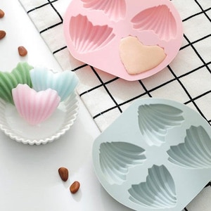 May include: Two silicone molds for making heart-shaped candies. One mold is pink and has a shell design. The other mold is light blue and has a shell design.