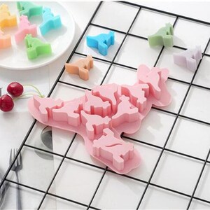 May include: Pink silicone mold with multiple dinosaur shapes. The mold is on a black wire grid.