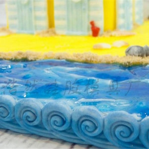 May include: A blue and white cake decorated to look like a beach scene. The cake has a sandy beach with a blue ocean and white waves. The ocean is decorated with blue frosting and the waves are made of white frosting.