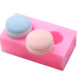 May include: Pink silicone mould with two cavities shaped like macarons. One cavity has a blue macaron and the other has a pink macaron.