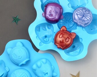 May include: Two blue silicone molds with cartoon faces, one mold has purple, red, and blue filled faces, the other mold is empty.