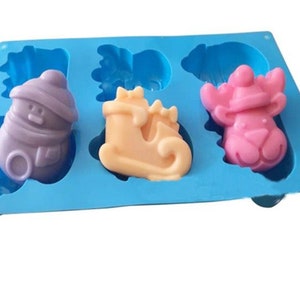 May include: A blue silicone mold with three Christmas-themed soap designs: a snowman, a sleigh, and a reindeer. The snowman is purple, the sleigh is beige, and the reindeer is pink.