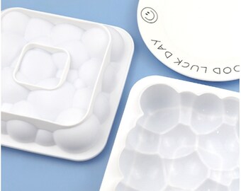 May include: Two white silicone moulds for making ice cubes. One mould is square with a large circle in the centre and the other mould has a honeycomb pattern. The moulds are on a light blue background.