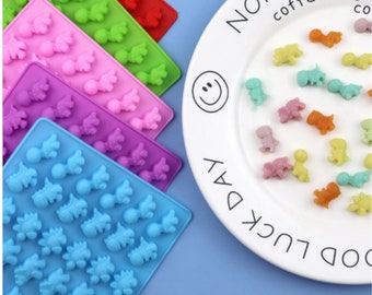 May include: Five silicone moulds for making candy or chocolate in the shape of dinosaurs. The moulds are in different colours: pink, green, purple, blue, and red. A plate of colourful dinosaur-shaped sweets is shown.