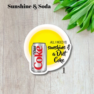 May include: A white sticker with a Diet Coke can illustration, set against a yellow sun design. The text reads "ALL I NEED IS sunshine & a Diet Coke." The sticker also has the text "Sunshine & Soda" at the top.