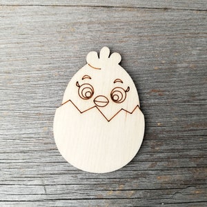 Peut inclure: Une découpe en bois d'un poussin éclosant d'un œuf. Le poussin a de grands yeux, un bec et une petite crête sur la tête.