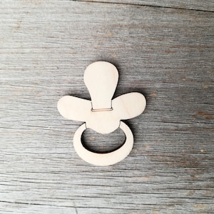 Puede incluir: Un recorte de madera de un chupete para bebé, un material de artesanía popular para decoraciones de baby shower o scrapbooking.