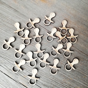 Puede incluir: Una colección de formas de chupete para bebés de madera, esparcidas sobre una superficie de madera marrón claro. Los chupetes son todos del mismo tamaño y forma, y están hechos de madera sin terminar.
