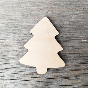 Pode incluir: Um enfeite de árvore de Natal de madeira simples, em forma de pinheiro tradicional, com uma superfície lisa de cor castanho claro. O enfeite está sobre um fundo de madeira cinzento.