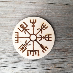 Op de afbeelding: Een ronde houten onderzetter met een gegraveerd Vegvisir-symbool, een Noors directioneel symbool, in het midden.