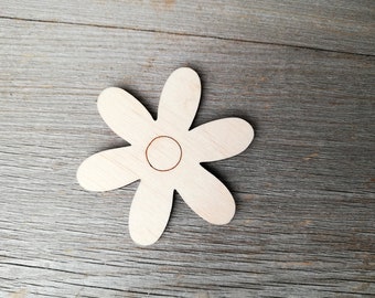 Puede incluir: Un recorte de flor de madera con seis pétalos y un círculo central. La flor está hecha de madera de color claro y tiene una estética simple y natural. El recorte se coloca sobre una superficie de madera gris y envejecida.