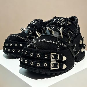 May include: Black platform shoes featuring silver metal spikes and buckles. The shoes have a chunky sole, a textured fabric upper, and multiple eyelets. The design is bold and edgy, suitable for alternative fashion.