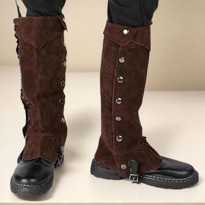 May include: Brown suede and leather spats with silver-tone button and buckle details. The spats cover the lower leg and foot, and are worn over black shoes. The spats have a vintage or historical aesthetic.