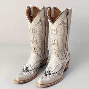 Op de afbeelding: Een paar off-white cowboylaarzen met ingewikkelde borduursels. De laarzen hebben een puntige neus, een blokhak en decoratieve studs. De hoge schacht is versierd met gedetailleerde stiksels, wat een klassieke Western-esthetiek creëert. Hoogte: ongeveer 30 cm.