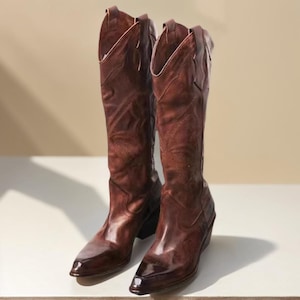 Op de afbeelding: Een paar kniehoge cowboylaarzen van bruin leer met een puntige neus, een blokhak en decoratieve, ton-sur-ton stikselpatronen op de schacht. De laarzen hebben een verweerde afwerking met variaties in de kleur van het leer.