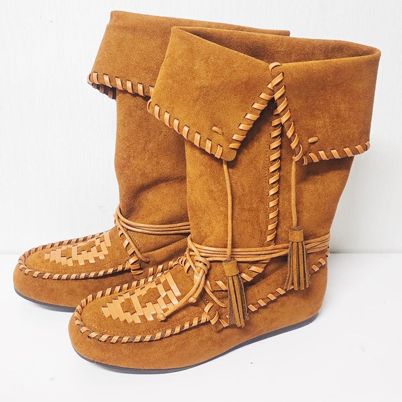 May include: A pair of brown suede boots with a folded-over top. The boots feature intricate leather stitching and woven details on the toe. Tassels and leather ties add decorative accents. The boots have a moccasin-style design.