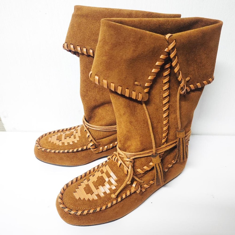 May include: A pair of brown suede boots with a folded-over top. The boots feature intricate leather stitching and a woven design on the toe. Tassels and laces add detail to the design.