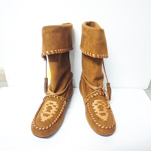 May include: A pair of brown suede boots with a moccasin-style design. The boots have a folded-over top, leather trim, and decorative stitching. Tassels hang from the top of the boots. The boots are displayed on a white surface.