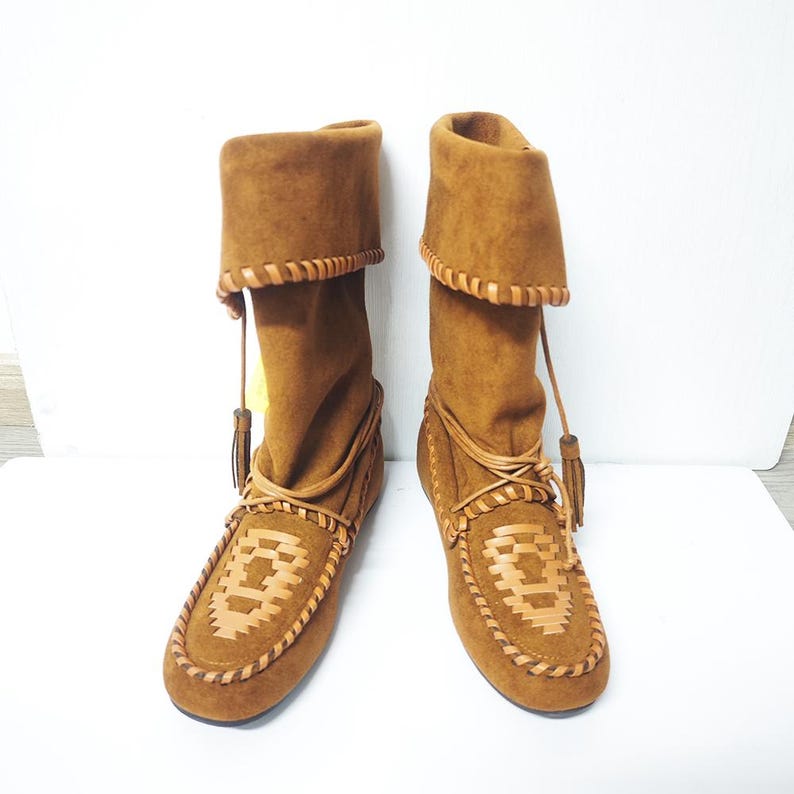 May include: A pair of brown suede boots with a moccasin-style design. The boots have a folded-over top, leather trim, and decorative stitching. Tassels hang from the top of the boots. The boots are displayed on a white surface.