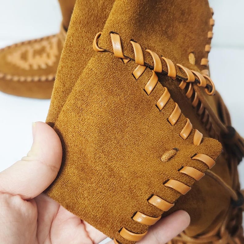 May include: Close-up of brown suede boots with leather stitching and fringe detail. The boots have a moccasin style with a soft, textured appearance. The stitching is a lighter shade of brown.