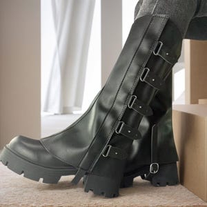 May include: Black knee-high boots with a chunky sole and multiple buckle fastenings. The boots have a faux leather appearance and a rounded toe. The heel is approximately 5 cm high. The boots are designed with a modern, edgy aesthetic.