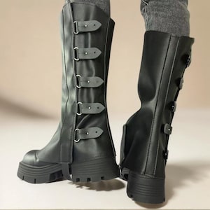 Puede incluir: Botas negras hasta la rodilla con múltiples cierres de hebilla y suela gruesa. Las botas tienen un acabado mate y un diseño robusto y atrevido. Las botas están hechas de material sintético.