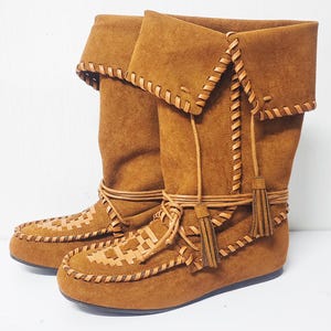 May include: A pair of brown suede boots with a folded-over top. The boots feature intricate leather stitching and woven details on the toe. Tassels and leather ties add decorative accents to the mid-calf boots.