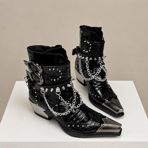 Op de afbeelding: Een paar zwarte lakleren enkellaarsjes met een krokodillenleertextuur, voorzien van zilverkleurige metalen neuzen, puntige studs en decoratieve kettingdetails. De laarsjes hebben verstelbare gespriemen en een blokhak.