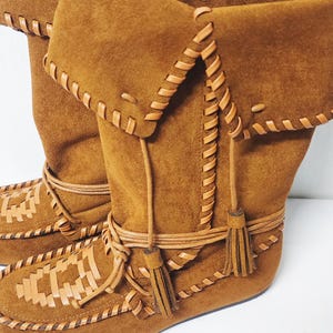 May include: Brown suede boots with a folded-over top. The boots feature decorative stitching, woven leather accents, and tassels. The boots have a moccasin-style design.