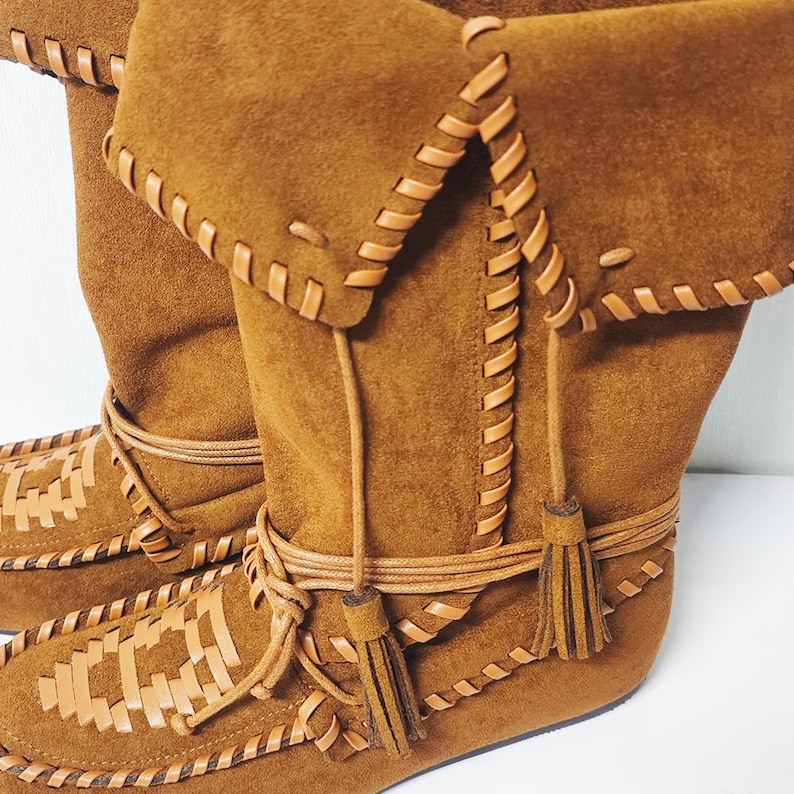 May include: Brown suede boots with a folded-over top. The boots feature decorative stitching, woven leather accents, and tassels. The boots have a moccasin-style design.