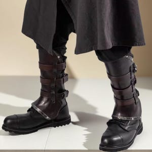May include: Black leather boots with multiple brown leather straps and silver-toned metal accents. The boots have a round toe and a thick sole. The upper portion of the boots is visible, with the lower part of the legs and clothing above.
