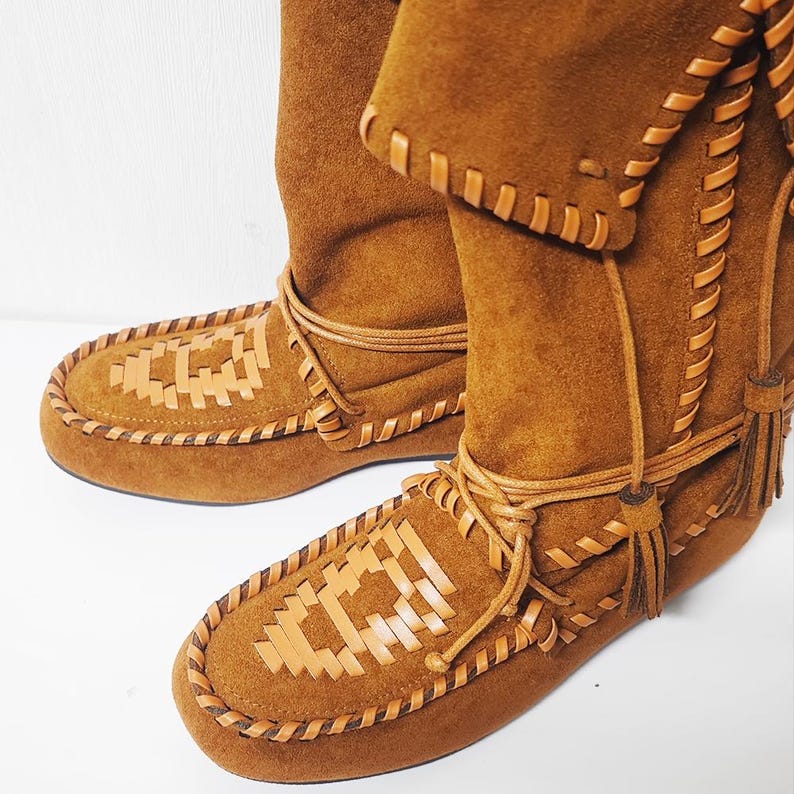 May include: Brown suede moccasin boots with decorative stitching and woven leather accents on the toe. The boots have a tall shaft and leather ties with tassels. The footwear is designed for comfort and style.