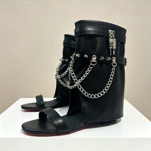 May include: Black leather sandal boots with silver chain and stud accents. The boots have a flat sole with a red bottom. A silver padlock detail is visible on the upper shaft.