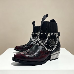 Pode incluir: Um par de botins pretos e bordô com detalhes em metal prateado. As botas apresentam um design de chama na biqueira, uma corrente e uma tira com tachas, e uma presilha. O salto é prateado e as botas têm bico fino.