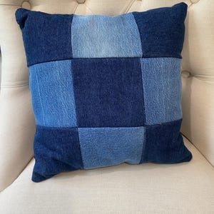 May include: A square throw pillow constructed from a patchwork of denim fabric squares. The design features an alternating grid pattern of dark indigo blue and lighter faded blue denim patches, creating a textured, multi-tonal blue appearance.