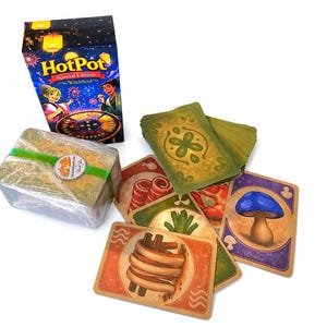 May include: A board game box titled "HotPot Special Edition" with a vibrant illustration. A stack of cards is wrapped in plastic and tied with green ribbon. Several illustrated game cards are scattered around.