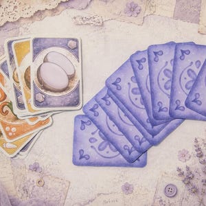 Pode incluir: Um baralho de cartas retangulares com versos roxos de padrão floral e faces que exibem ilustrações de vários alimentos, incluindo um coco, dispostas em leque sobre uma superfície roxa clara com detalhes em renda.