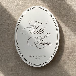 May include: Oval table number card with the words "Table Seven" in elegant script. The card also includes the names "Megan & Hunter" and the date "6.13.26" in a smaller font. The card has a black border.