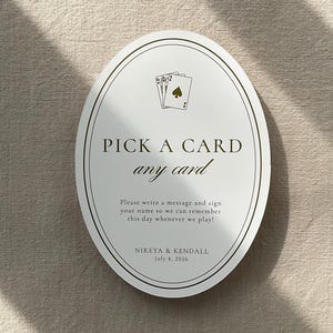 May include: An oval-shaped card with the text "PICK A CARD any card" in a serif font. The card features a playing card illustration and the names "NIKEYA & KENDALL" with the date "July 4, 2026". The card is on a neutral background.