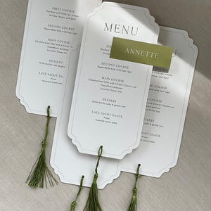 May include: Elegant white menu cards with a scalloped edge, featuring a detailed menu. Each card is adorned with a green tassel. A name card with the name "Annette" is also present.
