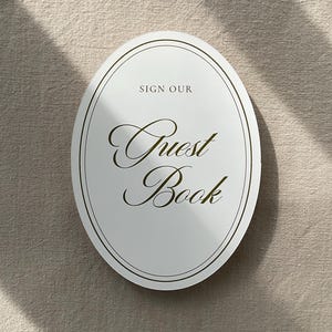 May include: An oval-shaped guest book with the words "SIGN OUR" above and "Guest Book" in elegant script. The book is white with a thin gold border and text, set against a neutral background.