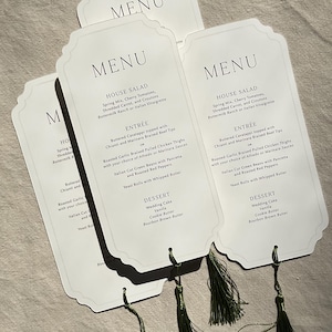 May include: White menu cards with a decorative top edge and green tassels. The cards display a menu with headings like "HOUSE SALAD" and "ENTRÉE". The menu includes items such as "Wedding Cake" and "Roasted Garlic Braised Pulled Chicken Thighs".