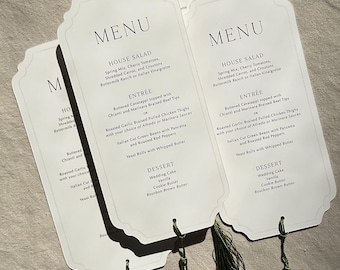 Könnte beinhalten: Ein Set aus drei cremeweißen Menükarten mit gewelltem Rand und schwarzem Aufdruck, die Optionen für Salat, Hauptgericht und Dessert auflisten. Jede Karte hat eine dunkelgrüne Zierquaste, die mit einer dünnen Kordel unten befestigt ist.