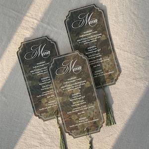 May include: Three menu cards with a floral design and the word "Menu" in elegant script. Each card has a green tassel. The menu items are listed in a decorative font. The cards are on a neutral-coloured surface.