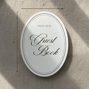 May include: White oval guest book sign, 12.7 cm wide and 17.8 cm tall, with elegant script that reads "Guest Book". The sign has a double border and the words "Sign Our" above the script.