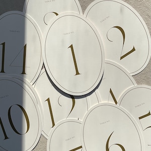 May include: A collection of ivory oval table number cards with gold trim and numbering. Each card displays a different number in a bold, gold font, ranging from 1 to 14. The words "TABLE NO." are printed at the top of each card.