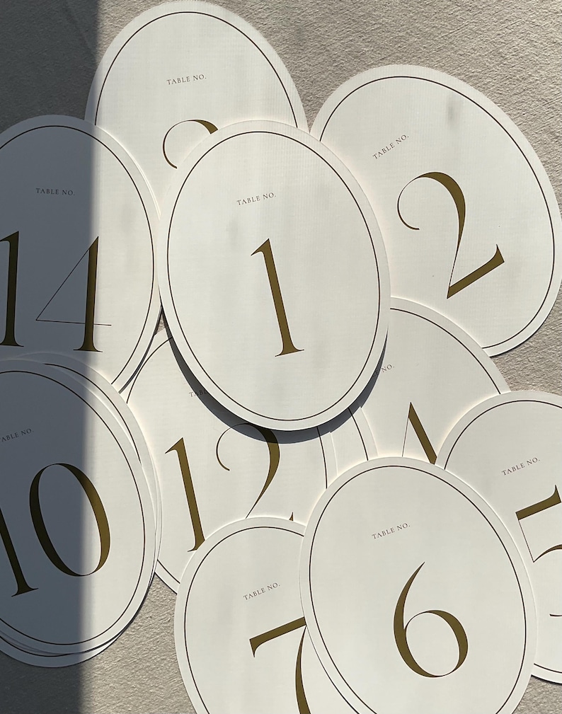 May include: A collection of ivory oval table number cards with gold trim and numbering. Each card displays a different number in a bold, gold font, ranging from 1 to 14. The words "TABLE NO." are printed at the top of each card.
