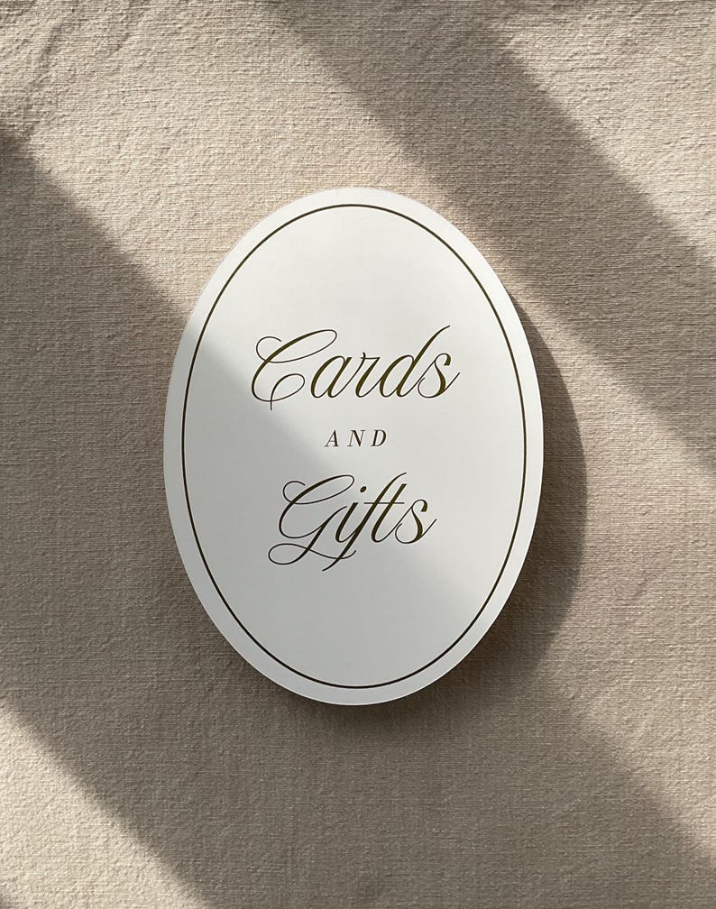 May include: An oval, white sign with the words "Cards and Gifts" in elegant, gold-colored script. The sign has a thin, dark border and is set against a neutral, textured background, with shadows adding depth.